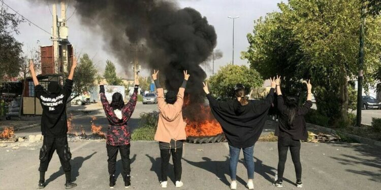 Five young protestors stand in the middle of the street in Saqqez with their hands in the air in front of a tire up in smoke and flames.